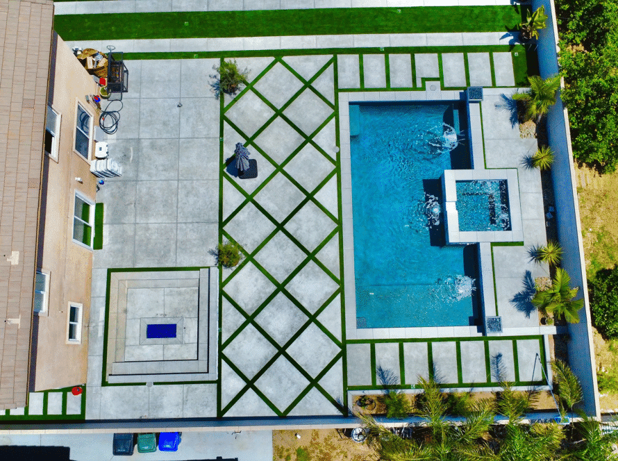 Aerial view of a backyard pool and spa with geometric concrete decking, artificial grass accents, and landscaped areas.