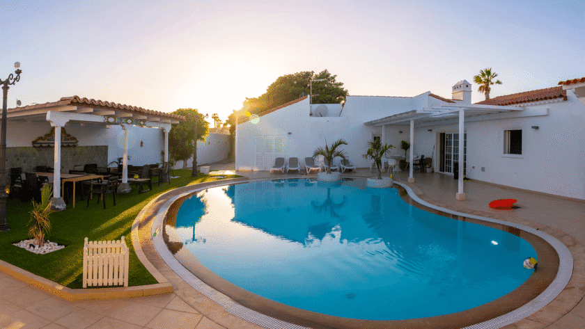 Resort-style backyard swimming pool surrounded by white stucco buildings, covered outdoor seating, lounge chairs, and landscaped areas during sunset.