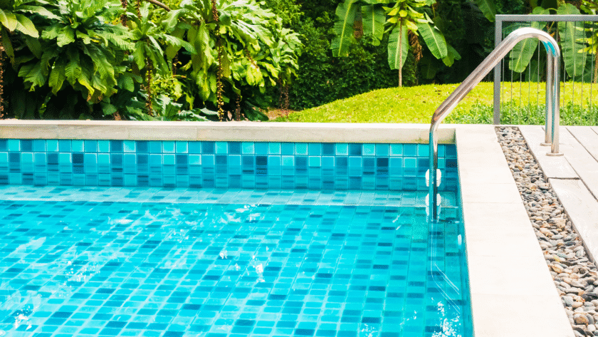 Close-up view of a bright blue tiled swimming pool with a stainless-steel handrail, light stone coping, and lush tropical landscaping in the background.