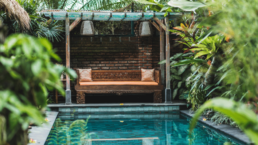 Tropical poolside retreat with a rustic wooden lounge pavilion, ornate bench seating, lush greenery, and a serene swimming pool.