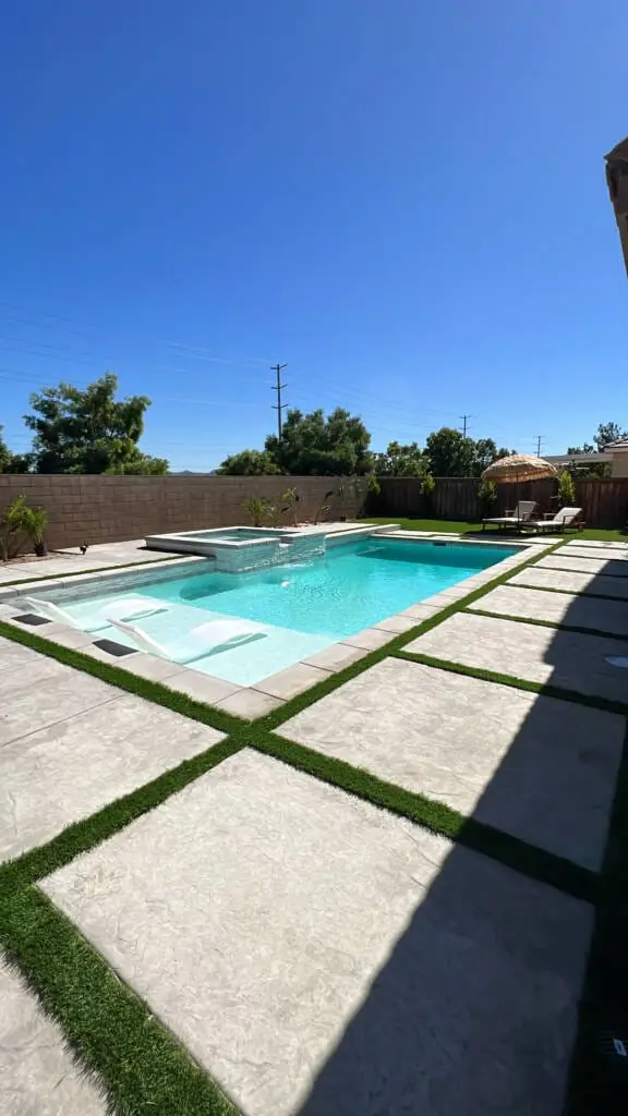 Modern backyard swimming pool with raised spa, shallow tanning ledge, geometric concrete patio, and artificial grass landscaping.