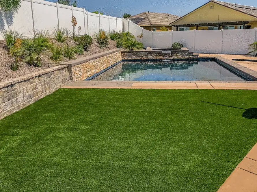Backyard swimming pool with a raised stone waterfall feature, landscaped rock garden, and artificial grass lawn.